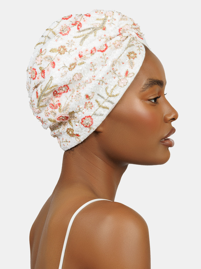 Profile view of black female model wearing hand made sequin-embellished white turban with a delicate red and pink flower pattern designed by Maryjane Claverol